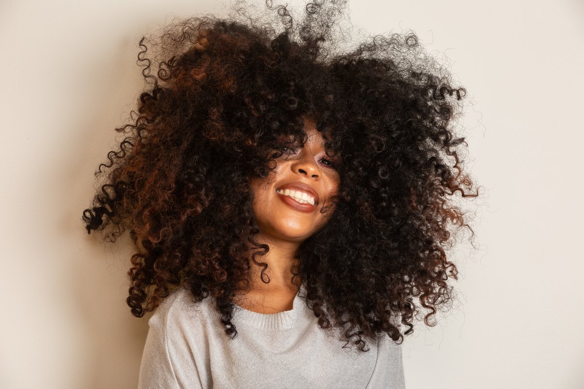 A portrait of a smiling woman with a large curly afro on a plain cream studio background.